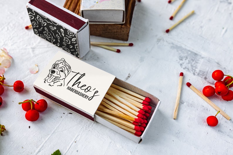 May include: An open matchbox with the text "Theo's Haberdashery" and a dog illustration. The matches have red tips. Other matchboxes are in the background. Red berries and matches are scattered around the matchbox.