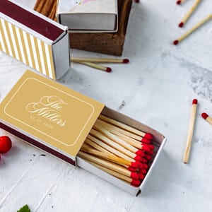 May include: A rectangular matchbox with a gold and white striped cover and a second open matchbox featuring a gold label with the white text The Millers and the date 08.07.2026. The open box contains wooden matches with red tips, surrounded by loose matches and red berries.
