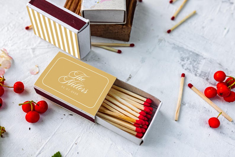 May include: A rectangular matchbox with a gold and white striped cover and a second open matchbox featuring a gold label with the white text The Millers and the date 08.07.2026. The open box contains wooden matches with red tips, surrounded by loose matches and red berries.