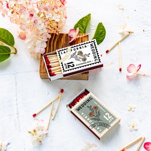 May include: Two rectangular matchboxes with red-tipped matches, featuring a stamp-like design with the text "California Wedding", "San Francisco", "12 Apr", and "Ann + Patrick" alongside a floral illustration in black and white or sage green.