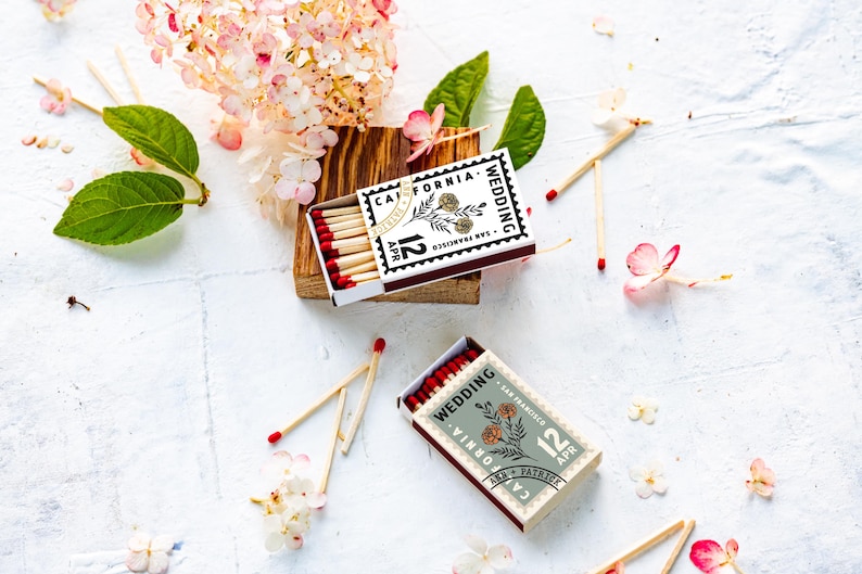 May include: Two rectangular matchboxes with red-tipped matches, featuring a stamp-like design with the text "California Wedding", "San Francisco", "12 Apr", and "Ann + Patrick" alongside a floral illustration in black and white or sage green.