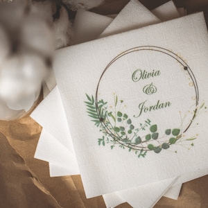 May include: A stack of white linen napkins with a printed design. The design features a gold-coloured circular frame with the names "Olivia & Jordan" in green script, surrounded by green botanical illustrations.