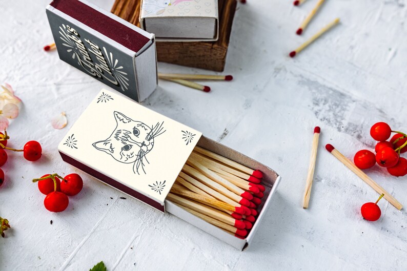 May include: A collection of matchboxes with a cat illustration on one. The matches have red tips. Other matchboxes have decorative designs. Red berries and matches are scattered on a white and gray surface.