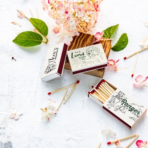 May include: Matchboxes with illustrations and text, including "Theo's", "Bar Luna", and "Ranger Station". The matchboxes are white with maroon accents and contain wooden matches with red tips. Floral and leaf accents surround the matchboxes.