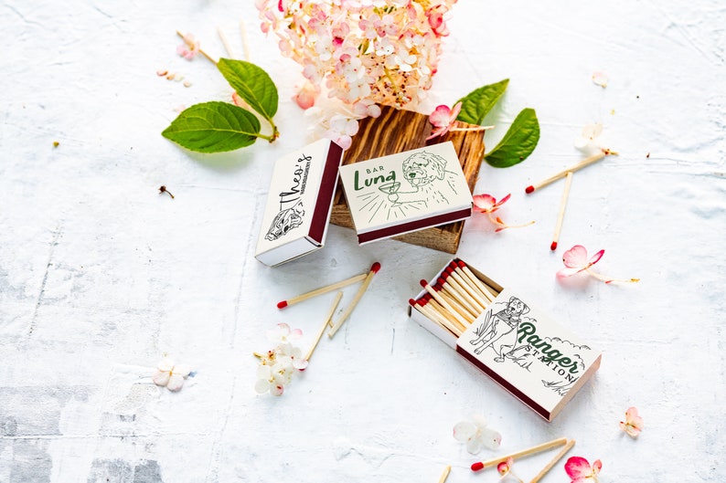 May include: Matchboxes with illustrations and text, including "Theo's", "Bar Luna", and "Ranger Station". The matchboxes are white with maroon accents and contain wooden matches with red tips. Floral and leaf accents surround the matchboxes.