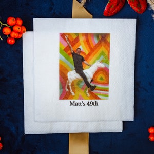 K&ouml;nnte beinhalten: Wei&szlig;e quadratische Servietten mit einem farbenfrohen Bild einer Person, die ein Einhorn reitet, mit dem Text "Matt's 49th". Der Hintergrund ist ein Regenbogen-Design. Die Servietten sind auf einer dunkelblauen Oberfl&auml;che mit dekorativen Elementen gestapelt.