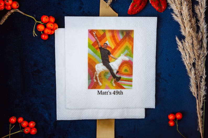 K&ouml;nnte beinhalten: Wei&szlig;e quadratische Servietten mit einem farbenfrohen Bild einer Person, die ein Einhorn reitet, mit dem Text "Matt's 49th". Der Hintergrund ist ein Regenbogen-Design. Die Servietten sind auf einer dunkelblauen Oberfl&auml;che mit dekorativen Elementen gestapelt.