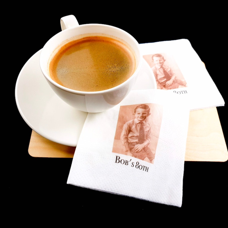 K&ouml;nnte beinhalten: Eine wei&szlig;e Kaffeetasse mit dunklem Kaffee steht auf einer passenden Untertasse. Zwei wei&szlig;e Servietten mit einem sepiafarbenen Foto eines jungen Jungen und dem Text "Bob's 80th" liegen neben der Tasse. Die Gegenst&auml;nde liegen auf einer hellen Holzoberfl&auml;che.