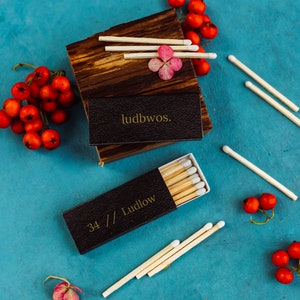 May include: Two boxes of matches on a blue background. The boxes are black with gold lettering. One box says "34 // Ludlow" and the other says "ludbws."