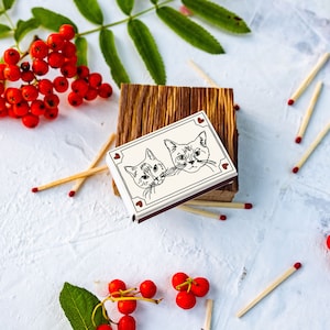 May include: A matchbox with a cat illustration, resting on a wooden block, surrounded by matches and red berries. The matchbox features two cat faces with heart accents. The matches have red tips.