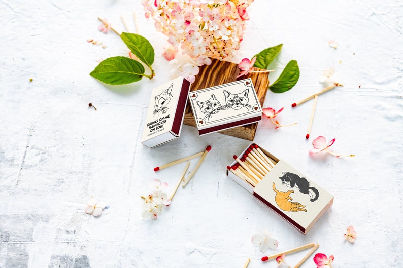 May include: Matchboxes with cat illustrations and the text "DRINKS ON ME, HANGOVER ON YOU!". The matchboxes are cream-colored with maroon accents and contain wooden matches with red tips. The image includes flowers and leaves.