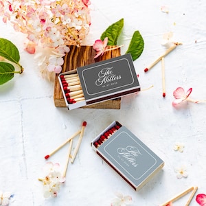 May include: Two rectangular matchboxes with dark gray covers featuring the white text The Millers and a date. Each box contains wooden matches with red tips. The boxes are arranged with loose matches and scattered pink and white flower petals.