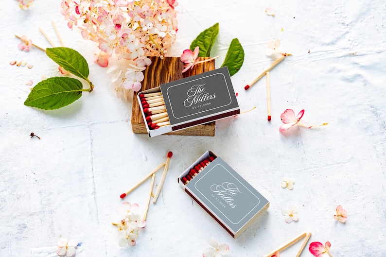 May include: Two rectangular matchboxes with dark gray covers featuring the white text The Millers and a date. Each box contains wooden matches with red tips. The boxes are arranged with loose matches and scattered pink and white flower petals.