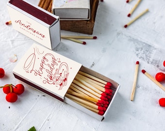 Puede incluir: Cajas de cerillas con cerillas de punta roja, una abierta y otra cerrada. La caja abierta muestra el texto "Happy Holidays" en escritura roja. La caja cerrada dice "The Andersons Christmas 2025". También se ven bayas rojas y cerillas dispersas.