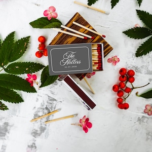 May include: A gray matchbox with the white text The Millers and 05.07.2020 printed on the front. The box is open, revealing wooden matches with red tips. Several loose matches, green leaves, and clusters of small red berries surround the box.