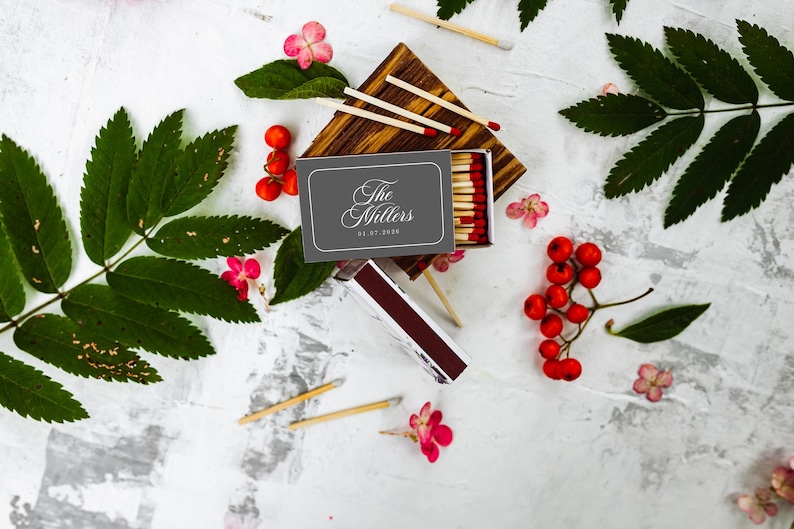 May include: A gray matchbox with the white text The Millers and 05.07.2020 printed on the front. The box is open, revealing wooden matches with red tips. Several loose matches, green leaves, and clusters of small red berries surround the box.