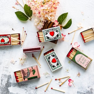May include: Assortment of matchboxes with various designs and colors. The matchboxes feature illustrations of hearts, a bulldog, a bird, and text in French and English. The matchboxes are scattered on a white surface with pink flowers.