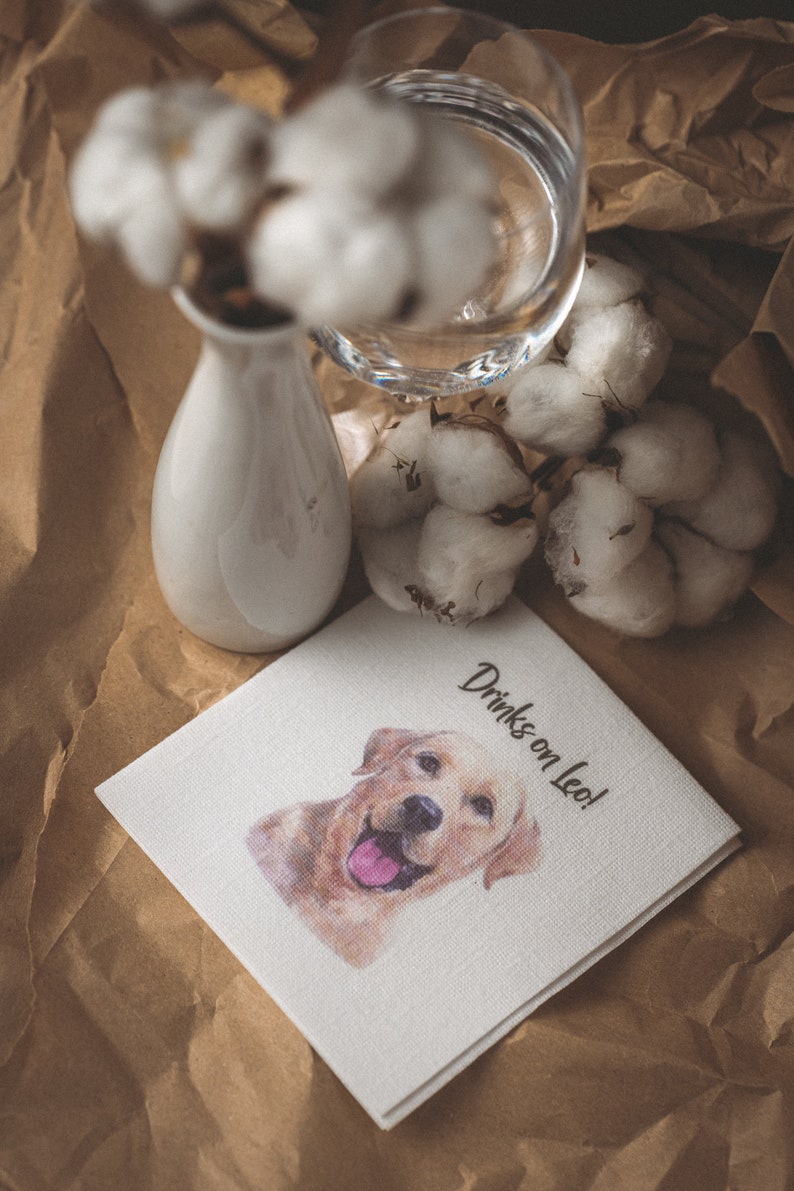 May include: A white linen cocktail napkin featuring a watercolour image of a golden retriever. The napkin has the text "Drinks on Leo!" in black script. The napkin is on a brown paper surface.