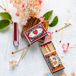 May include: Two boxes of matches, one red with a heart and the text "The Sissons Est. 2021 Made in USA Safety Matches", and the other orange with a bulldog and the text "Classic CTP True Tattoo".  The boxes are surrounded by pink and white flowers and wooden matches.