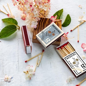 May include: Two rectangular matchboxes with dark maroon edges and white tops featuring a black stamp-style design that reads Wedding, California, San Francisco, 12 Apr, and Ann + Patrick, with a floral illustration and red-tipped wooden matches.