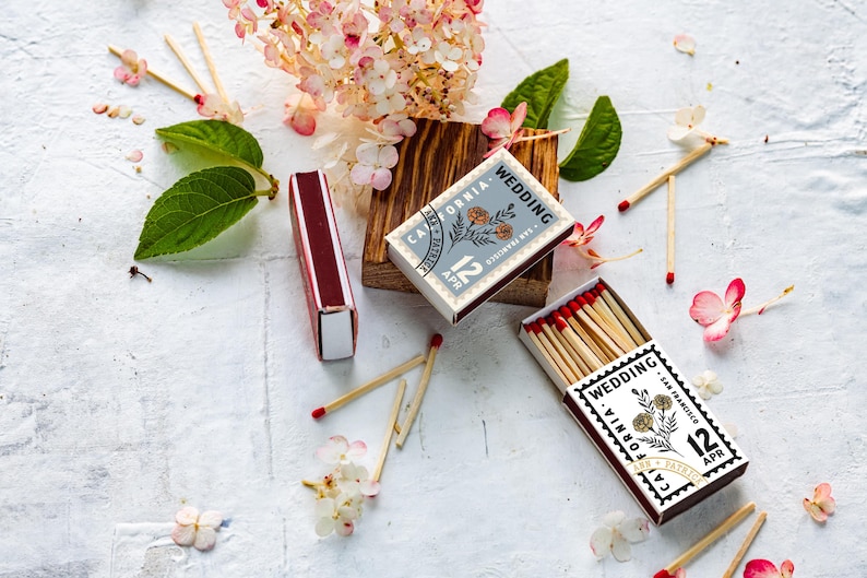 May include: Two rectangular matchboxes with dark maroon edges and white tops featuring a black stamp-style design that reads Wedding, California, San Francisco, 12 Apr, and Ann + Patrick, with a floral illustration and red-tipped wooden matches.