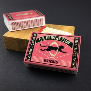 May include: Two rectangular matchboxes with a pink background and black borders. The front features a black panther jumping through a yellow ring of fire, with the text ILB DRIVERS CLUB, ESSENTIAL EQUIPMENT, and MATCHES printed in black.