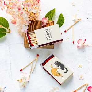 May include: Two matchboxes with red-tipped matches are displayed. One box has the text "The Emmans, March 22, 2024". The other box features illustrations of two cats. The scene is decorated with flowers and leaves.