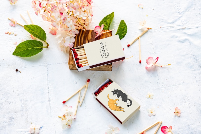 May include: Two matchboxes with red-tipped matches are displayed. One box has the text "The Emmans, March 22, 2024". The other box features illustrations of two cats. The scene is decorated with flowers and leaves.