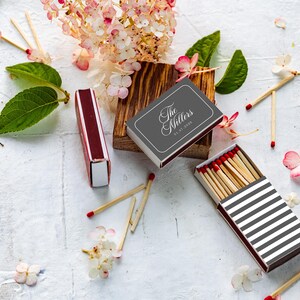 May include: A set of matchboxes featuring a dark gray box with the white text The Millers and a date, a maroon box, and a box with black and white horizontal stripes containing wooden matches with red tips, surrounded by pink hydrangea flowers and green leaves.