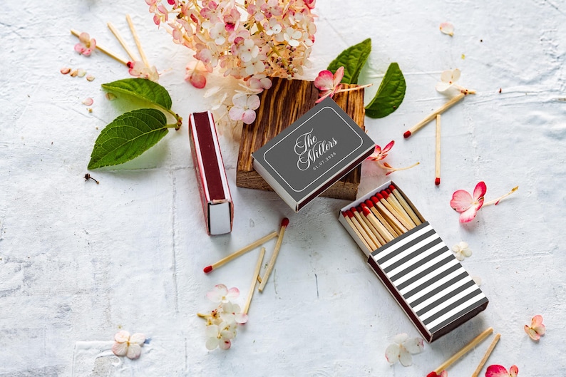 May include: A set of matchboxes featuring a dark gray box with the white text The Millers and a date, a maroon box, and a box with black and white horizontal stripes containing wooden matches with red tips, surrounded by pink hydrangea flowers and green leaves.