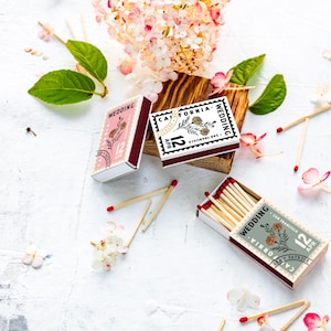 May include: Three rectangular matchboxes featuring a stamp-like design with the text Wedding, California, and 12 Apr. The boxes are colored in pink, white, and sage green, each decorated with a floral illustration. Several wooden matches are scattered.