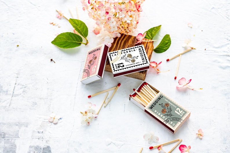 May include: Three rectangular matchboxes featuring a stamp-like design with the text Wedding, California, and 12 Apr. The boxes are colored in pink, white, and sage green, each decorated with a floral illustration. Several wooden matches are scattered.