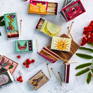 May include: Assortment of colorful matchboxes with various designs, including a butterfly, a sun, a lemon, and a woman on a swing. The matchboxes are arranged on a white surface with a green leaf and red berries.