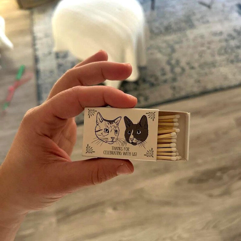 May include: A small, open matchbox held in a hand. The box features illustrations of two cats and the text "THANKS FOR CELEBRATING WITH US!". The matchsticks are visible inside the box.