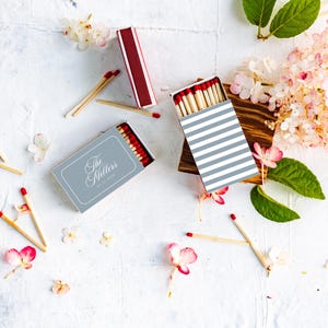 May include: Two rectangular matchboxes with red-tipped wooden matches. One box features a grey cover with the text The Millers, while the other has a white and grey striped pattern. Scattered matches and pink hydrangea flowers surround the boxes.