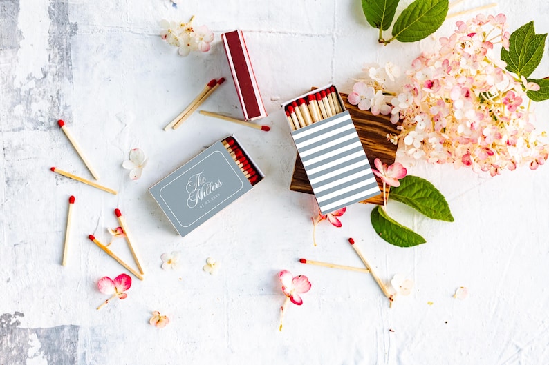 May include: Two rectangular matchboxes with red-tipped wooden matches. One box features a grey cover with the text The Millers, while the other has a white and grey striped pattern. Scattered matches and pink hydrangea flowers surround the boxes.
