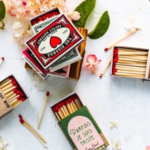 May include: Assortment of matchboxes with various designs.  The matchboxes feature illustrations of a heart, a bottle, a dog, and a phrase in French.  The matchboxes are on a white surface with pink flowers.