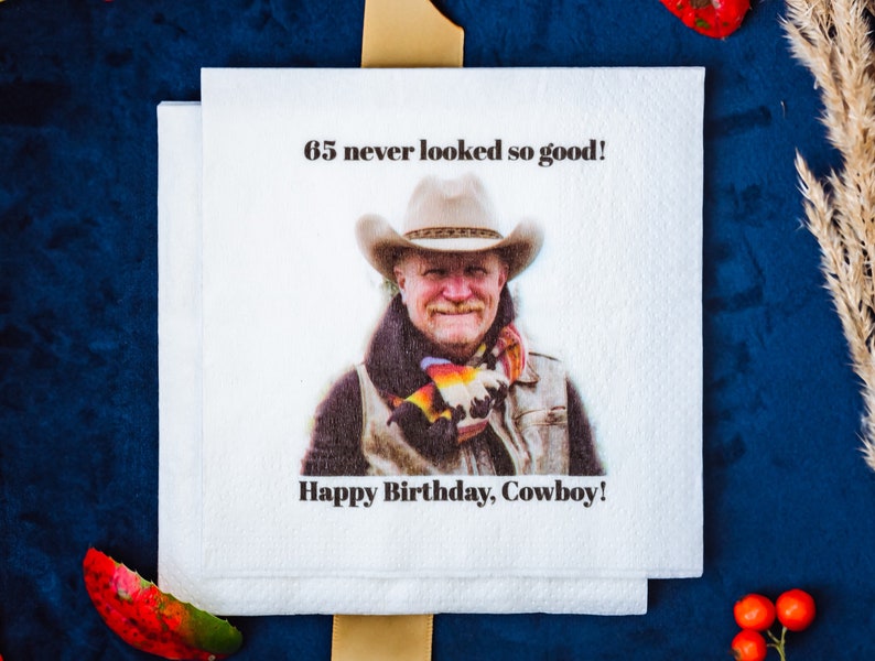 K&ouml;nnte beinhalten: Wei&szlig;e Papierservietten mit einem aufgedruckten Bild eines Mannes mit Cowboyhut. Der Text "65 never looked so good!" und "Happy Birthday, Cowboy!" sind auf den Servietten aufgedruckt. Die Servietten sind auf einer blauen Oberfl&auml;che gestapelt.