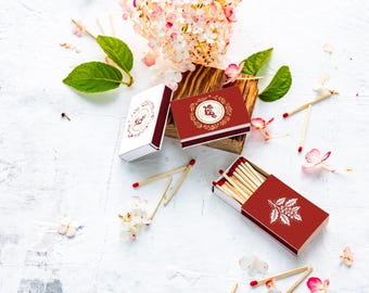 Puede incluir: Vista aérea de tres cajas de cerillas, una blanca y dos rojas, con detalles florales y de hojas. La caja blanca tiene la palabra "Love" en una fuente decorativa. Cerillas y flores dispersas.