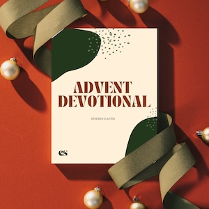 May include: A cream-colored card with green abstract shapes and a title that reads "Advent Devotional" with the author's name "Sherin Eapen" below. The card is surrounded by gold ornaments and green ribbon on a red background.