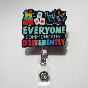 May include: A black and white badge holder with a colorful design and the text "Everyone Communicates Differently". The design includes a building block, a hand with a rock and roll sign, and crayons.