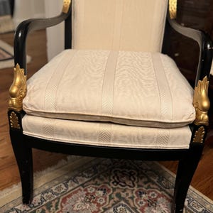 Puede incluir: Sill&oacute;n antiguo con estructura negra y detalles dorados. El asiento y el respaldo son de color crema con un patr&oacute;n de rayas verticales. Los reposabrazos son curvos y las patas de la silla son rectas.