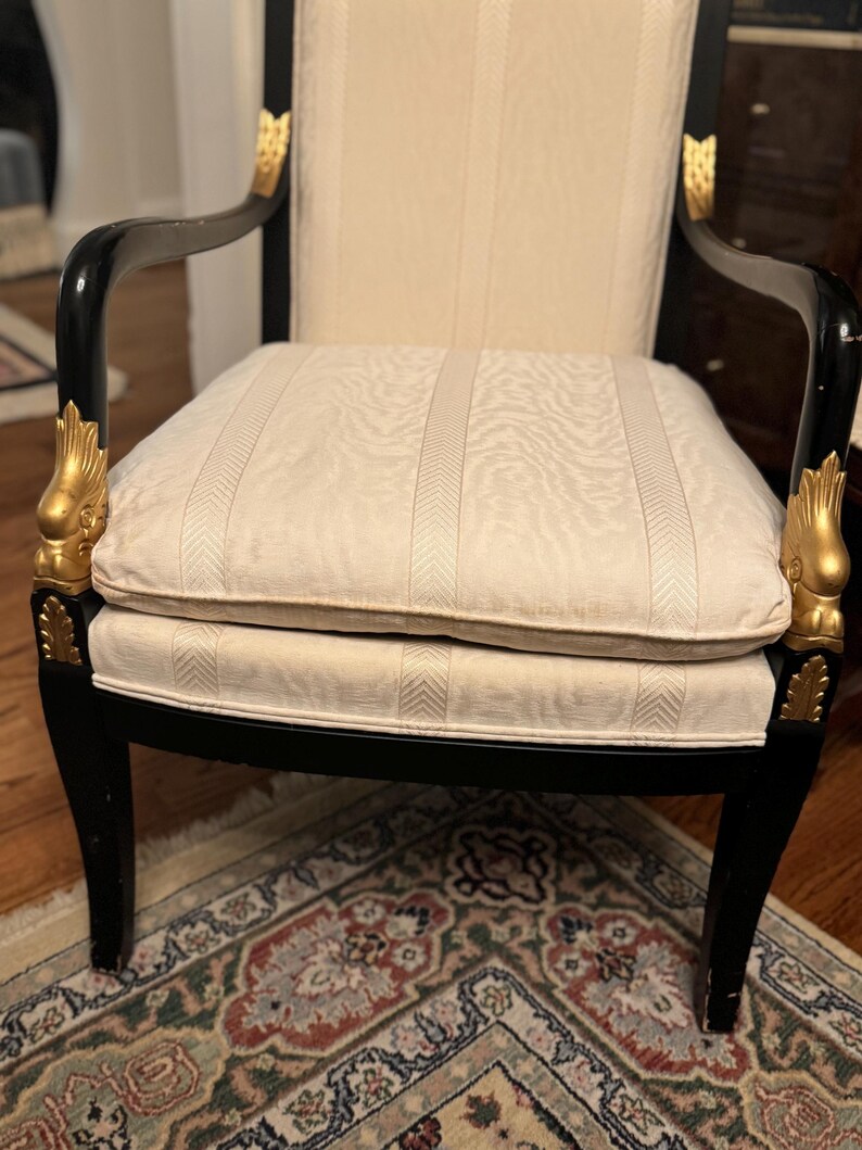 Puede incluir: Sill&oacute;n antiguo con estructura negra y detalles dorados. El asiento y el respaldo son de color crema con un patr&oacute;n de rayas verticales. Los reposabrazos son curvos y las patas de la silla son rectas.