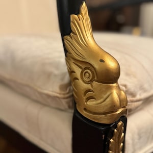 Puede incluir: Primer plano de un brazo de silla negro con detalles decorativos dorados. Los detalles dorados est&aacute;n tallados con un dise&ntilde;o de p&aacute;jaro. La silla est&aacute; sobre un coj&iacute;n crema.