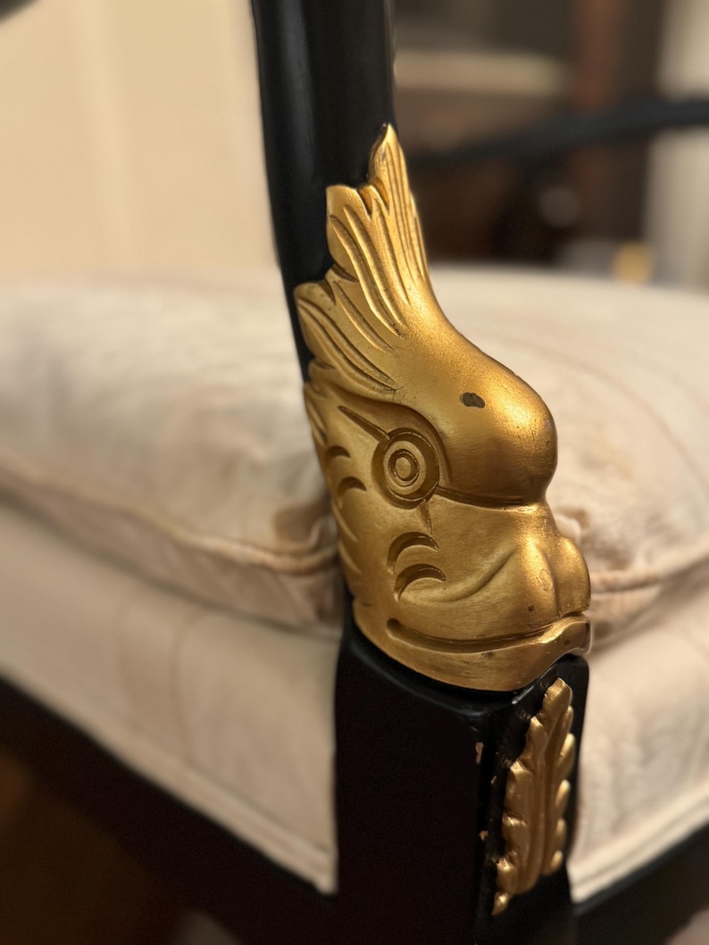 Puede incluir: Primer plano de un brazo de silla negro con detalles decorativos dorados. Los detalles dorados est&aacute;n tallados con un dise&ntilde;o de p&aacute;jaro. La silla est&aacute; sobre un coj&iacute;n crema.