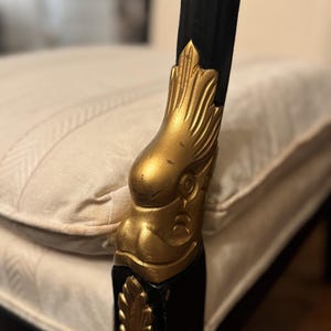 Puede incluir: Primer plano de un brazo de silla negro con un detalle de cabeza de &aacute;guila dorada. El brazo de la silla forma parte de un mueble m&aacute;s grande, con un coj&iacute;n de color crema visible en el fondo. La cabeza del &aacute;guila est&aacute; intrincadamente tallada.