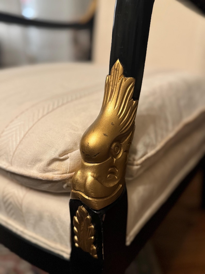 Puede incluir: Primer plano de un brazo de silla negro con un detalle de cabeza de &aacute;guila dorada. El brazo de la silla forma parte de un mueble m&aacute;s grande, con un coj&iacute;n de color crema visible en el fondo. La cabeza del &aacute;guila est&aacute; intrincadamente tallada.