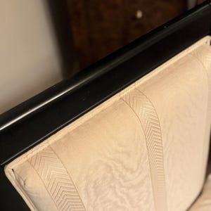 Puede incluir: Primer plano de un marco de silla de madera negra con un respaldo tapizado de color crema. El respaldo presenta paneles verticales con un sutil patr&oacute;n de espiga, creando un dise&ntilde;o texturizado. La silla parece ser parte de un conjunto.