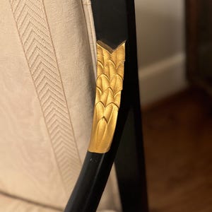 Puede incluir: Primer plano de un brazo de silla negro con un acento de hoja dorada. El brazo de la silla tiene un dise&ntilde;o decorativo de hoja dorada y se coloca contra una tela color crema con un sutil patr&oacute;n de espiga. La silla parece ser parte de un conjunto de muebles m&aacute;s grande.