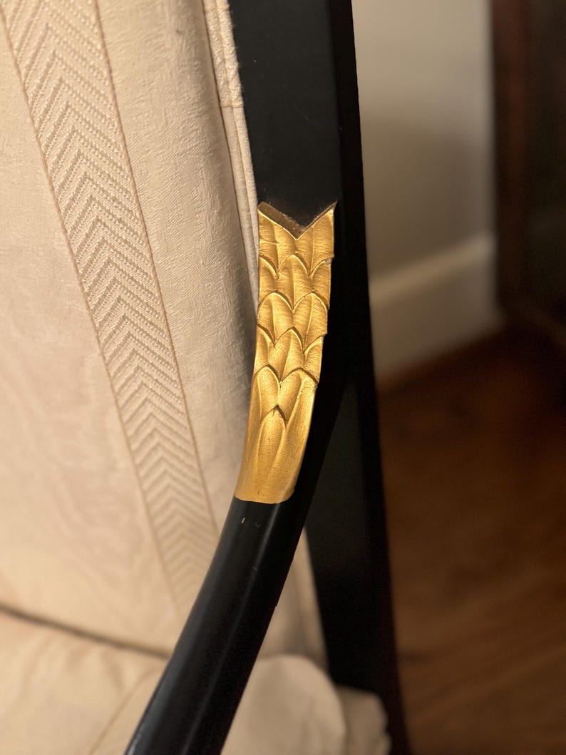 Puede incluir: Primer plano de un brazo de silla negro con un acento de hoja dorada. El brazo de la silla tiene un dise&ntilde;o decorativo de hoja dorada y se coloca contra una tela color crema con un sutil patr&oacute;n de espiga. La silla parece ser parte de un conjunto de muebles m&aacute;s grande.